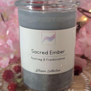 Op de afbeelding: Een grijze kaars in een heldere glazen pot met de tekst "Sacred Ember" en "Nutmeg & Frankincense" op een wit etiket. De kaars maakt deel uit van de "Blossom Collection". Roze bloemen op de achtergrond.