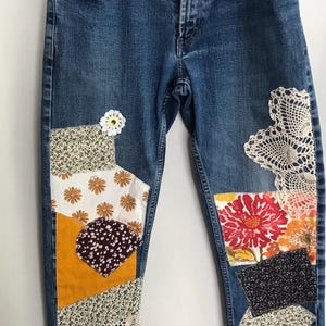 Peut inclure: Jean bleu avec des patchworks décoratifs. Les patchs présentent des tissus floraux, en velours côtelé et en dentelle de différentes couleurs. Un ornement blanc en forme de marguerite est près du haut de la jambe gauche. Le jean est suspendu avec une pince à linge en bois.