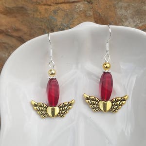 May include: A pair of dangle earrings with red glass beads and gold-toned angel wings. The earrings are on a white plate.