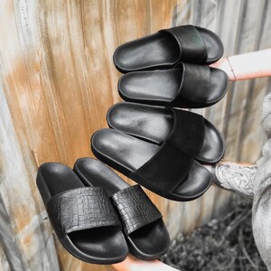 May include: Five pairs of black leather slides. The slides have a variety of textures and designs, including smooth leather, textured leather, and a crocodile pattern.