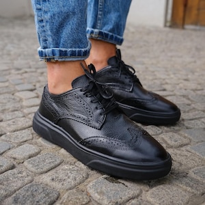 Op de afbeelding: Zwarte leren brogues met een witte zool en een vetersluiting. De schoenen hebben een geperforeerd ontwerp en worden gedragen met een blauwe jeans.