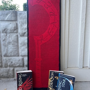 Op de afbeelding: Een grote, rode gehaakte wandhanger met een zwarte rand, met een gedetailleerd ontwerp. Drie boeken met de titels "Red Rising", "Golden Son" en "Morning Star" staan voor de wandhanger. De boeken zijn van Pierce Brown.