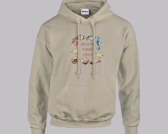 Könnte beinhalten: Ein beiger Hoodie mit einer Tasche auf der Vorderseite. Der Hoodie hat einen grafischen Druck von Muscheln und einem Seepferdchen mit dem Text "Beach Vibes Only".
