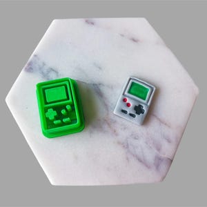 Op de afbeelding: Twee miniatuurvormige objecten in de vorm van een gameconsole. Eén is felgroen, de andere is grijs met rode en zwarte details. Beide hebben een scherm en knopdetails. Ze zijn geplaatst op een wit en grijs zeshoekig oppervlak.