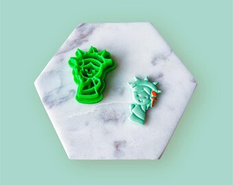 May include: Two green plastic cookie cutters shaped like the Statue of Liberty. One cutter is a detailed outline of the statue, while the other is a simplified version with a smiling face.