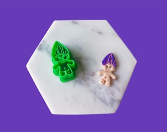May include: A green plastic troll-shaped cookie cutter and a small troll figurine with purple hair and a smiling face are displayed on a white marble hexagon against a purple background. The cookie cutter is in the shape of a troll.