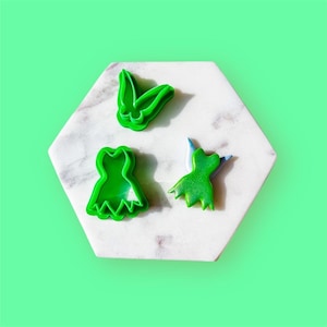 May include: Three green plastic cookie cutters shaped like a fairy, a dress, and a pair of wings.