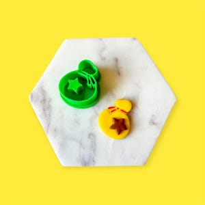 Op de afbeelding: Groene plastic koekjesvormer in de vorm van een geldzak met een ster erin. Ernaast ligt een gele fondantkoekje in de vorm van een geldzak met een bruine ster erin.
