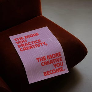 Pode incluir: Uma placa retangular rosa com texto vermelho que diz: "THE MORE YOU PRACTICE CREATIVITY, THE MORE CREATIVE YOU BECOME". A placa est&aacute; colocada em uma poltrona de veludo cotel&ecirc; marrom.