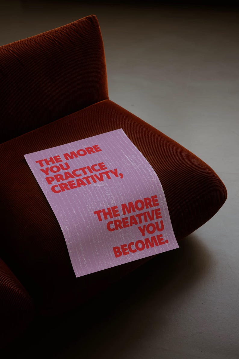 Pode incluir: Uma placa retangular rosa com texto vermelho que diz: "THE MORE YOU PRACTICE CREATIVITY, THE MORE CREATIVE YOU BECOME". A placa est&aacute; colocada em uma poltrona de veludo cotel&ecirc; marrom.