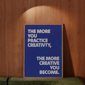 May include: A framed poster with the text "THE MORE YOU PRACTICE CREATIVITY, THE MORE CREATIVE YOU BECOME" in white letters on a blue background. The poster is set against a wooden wall and a green sofa.