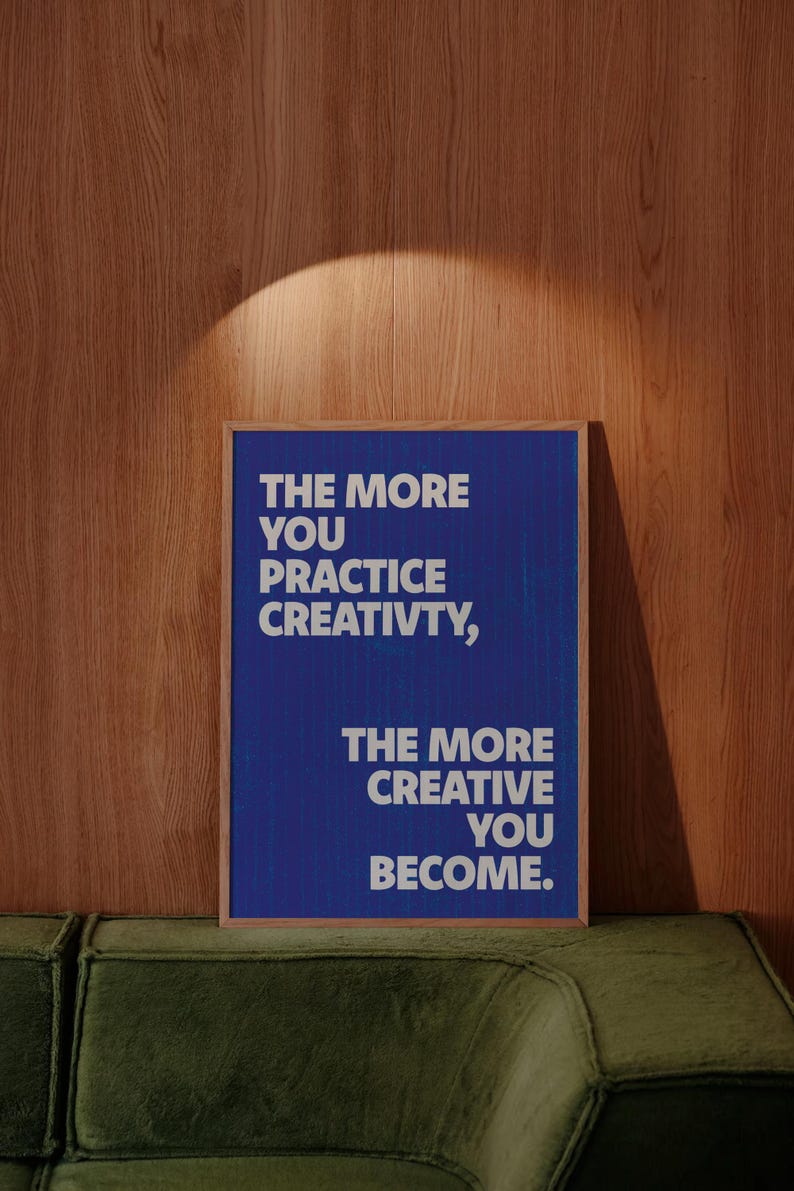 Pode incluir: Um poster emoldurado com o texto "THE MORE YOU PRACTICE CREATIVITY, THE MORE CREATIVE YOU BECOME" em letras brancas sobre um fundo azul. O poster est&aacute; colocado contra uma parede de madeira e um sof&aacute; verde.