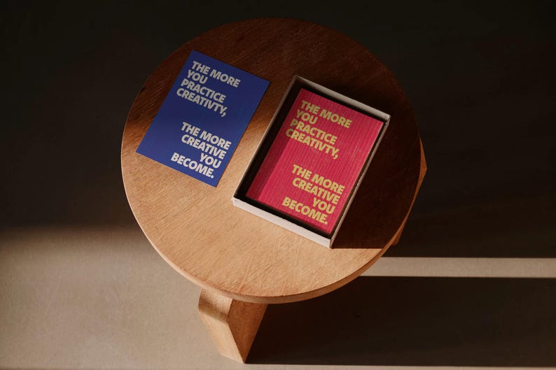 Pode incluir: Um cart&atilde;o azul e uma caixa de cart&otilde;es vermelhos com o texto "THE MORE YOU PRACTICE CREATIVITY, THE MORE CREATIVE YOU BECOME" numa mesa de madeira. O texto est&aacute; em ingl&ecirc;s.