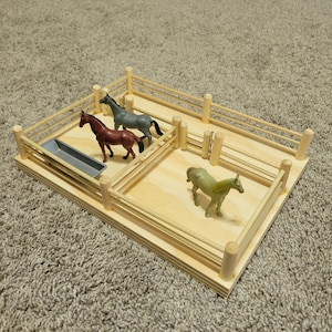 May include: A wooden horse stable with three toy horses in different colors: brown, gray, and green. The stable has a light wood base, a wooden fence, and a gray water trough. The toy horses are positioned inside the fenced areas.