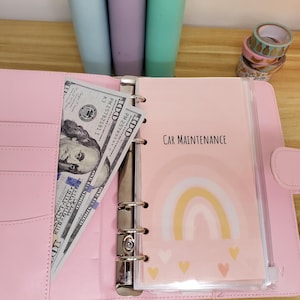 May include: A pink binder-style planner with a cash pocket holding several $100 bills. The planner has a section labeled "Car Maintenance" with a rainbow and heart design. Decorative tape rolls are visible in the background.