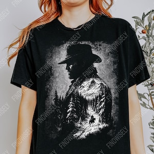 May include: Black t-shirt featuring a black and white graphic of a man in a hat, with a mountain and forest scene. The design includes a person on horseback. The shirt has a crew neck and short sleeves.