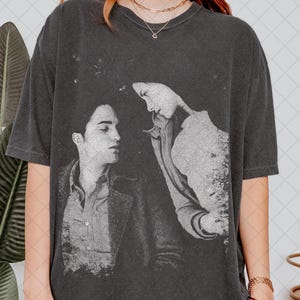 Könnte beinhalten: Ein anthrazitfarbenes Kurzarm-T-Shirt mit einem verwaschenen, einfarbigen Grafikdruck von zwei Personen, die sich gegenüberstehen. Das T-Shirt hat einen Rundhalsausschnitt und eine lockere Passform mit einer verblichenen Textur.
