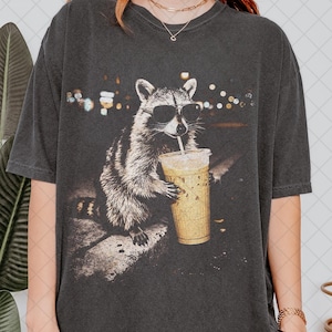 May include: A charcoal gray short-sleeve t-shirt featuring a graphic print of a raccoon wearing black sunglasses and drinking from a clear plastic cup with a straw. The raccoon is positioned on a concrete ledge against a dark, bokeh-lit background.