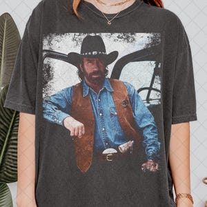 Puede incluir: Camiseta gris oscuro con una imagen desgastada de un hombre con sombrero de vaquero y camisa vaquera. La imagen muestra al hombre apoyado en un vehículo. La camiseta tiene un aspecto vintage y desgastado.