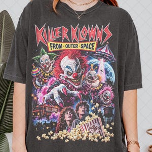 Op de afbeelding: Een antracietgrijs T-shirt met korte mouwen en een grafische print van de film Killer Klowns from Outer Space. Het ontwerp bevat meerdere clownfiguren, een ruimteschip, een emmer popcorn en de filmtitel in rode en gele tekst.