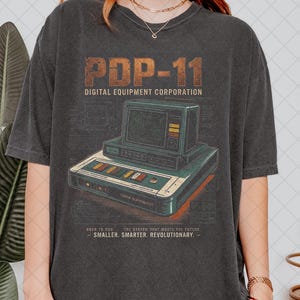 Może przedstawiać: Ciemnoszara koszulka z grafiką retro komputera. Projekt zawiera tekst "PDP-11 DIGITAL EQUIPMENT CORPORATION" oraz ilustrację komputera w stylu vintage. Tekst "BORN TO RUN - SMALLER. SMARTER. REVOLUTIONARY." jest również zawarty.