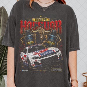 May include: A charcoal gray short-sleeve t-shirt featuring a graphic print of a race car, a bull, and two bottles labeled President 1994. The text Carson Hocevar is displayed above the bull, with NASCAR and Spire Motorsports logos below.