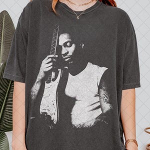 Puede incluir: Una camiseta de manga corta en color gris carbón con un gran estampado gráfico blanco desgastado de un músico sosteniendo una guitarra eléctrica. La prenda tiene cuello redondo y un corte holgado de estilo vintage.
