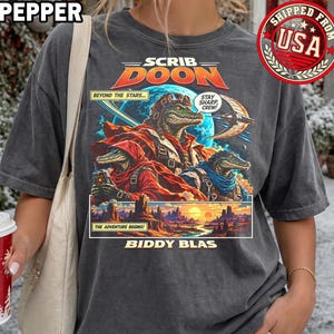 May include: A charcoal gray t-shirt featuring a graphic print of three anthropomorphic crocodiles in space gear. The text reads SCRIB DOON, BEYOND THE STARS, STAY SHARP CREW, THE ADVENTURE BEGINS, and BIDDY BLAS. The design includes a planet and a desert landscape.
