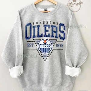 May include: A light gray sweatshirt with the text "EDMONTON OILERS" in blue, with a logo featuring a triangle and the words "OILERS" and "EST 1979". The sweatshirt has rolled-up white cuffs and is hanging on a wooden hanger.