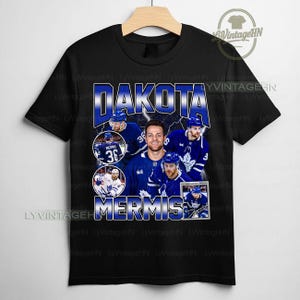 May include: Black t-shirt featuring a collage of images and the name "DAKOTA MERMIS" in blue and white. The number "36" is prominently displayed. The shirt showcases a hockey theme, with player images in blue and white.