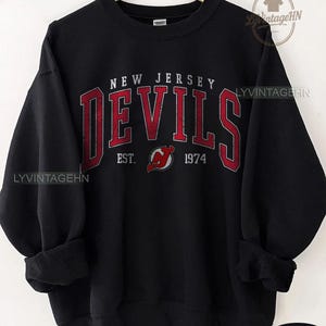 Może przedstawiać: Czarna bluza z napisem "NEW JERSEY DEVILS" w kolorze czerwonym, z wyeksponowanym słowem "DEVILS". Poniżej napis "EST. 1974" i logo. Bluza ma okrągły dekolt i długie rękawy.