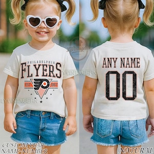 May include: A cream-coloured t-shirt with the words "PHILADELPHIA FLYERS" in black and orange, a team logo, and the year "1967". The back of the shirt shows "ANY NAME" above the number "00". The shirt is worn with blue denim shorts.