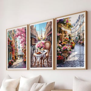 Può includere: Tre stampe artistiche incorniciate raffiguranti scene di strada parigine. Le stampe presentano colori vivaci, mostrando fiorerie, caffè e la Torre Eiffel. L'opera d'arte è incorniciata in legno chiaro ed è esposta sopra cuscini bianchi e un cesto intrecciato.