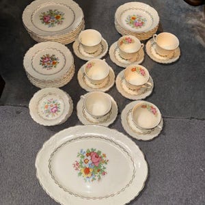 May include: A dinnerware set featuring off-white ceramic pieces with scalloped edges, gold trim, and a central floral bouquet pattern. The collection includes stacks of dinner plates, salad plates, bowls, and teacups with matching saucers.