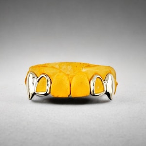 May include: A dental mold made of yellow material featuring metallic silver-colored caps on the two outer teeth on each side. The caps are shaped like pointed fangs, contrasting with the plain yellow teeth in the center of the dental impression.
