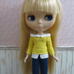 May include: A doll wearing a yellow crochet sweater with a white collar and cuffs, paired with dark blue crochet pants. The doll has long blonde hair and blue eyes. The outfit is handmade.