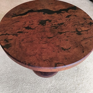May include: A circular wooden table with a reddish-brown burl wood top. The top has dark, organic patterns and a dark, linear inlay. The table has a pedestal base. The design is elegant and natural. The table's diameter is approximately 76 cm.