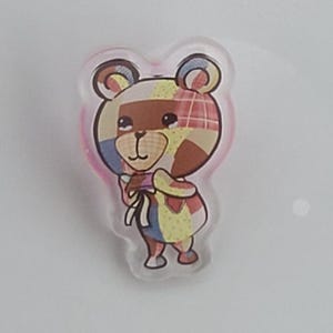 May include: A colorful bear-shaped pin with a patchwork design. The bear has a brown face and ears, and its body is made up of various colors and patterns, including blue, yellow, and red. The pin has a pink border.