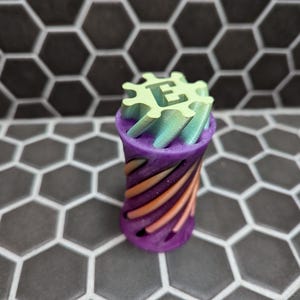 May include: A 3D-printed bottle stopper with a gear-shaped top and a cylindrical body. The top is light green with the letter "E". The body is purple with orange and teal stripes. The stopper is designed to fit into a bottle.