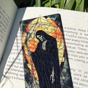 Op de afbeelding: Een glas-in-lood bladwijzer met een skeletfiguur in zwarte gewaden dat een zeis vasthoudt. De achtergrond is een levendige mix van oranje, gele en blauwe tinten. De bladwijzer heeft de titel "Fight Abate"