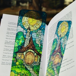 May include: A rectangular bookmark featuring a stained-glass style illustration of a small house with a round wooden door, a stone chimney, and a path, set against a bright yellow sun and green foliage, resting on an open book with printed text.