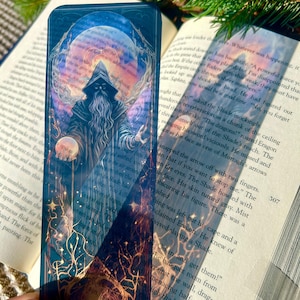 May include: A fantasy-themed bookmark featuring a wizard with a long beard and a staff, set against a backdrop of a moon and stars. The bookmark is rectangular and made of a transparent material.