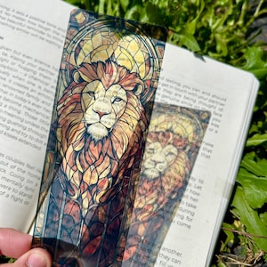 May include: A clear bookmark featuring a detailed illustration of a lion's head in warm tones of brown, orange, and yellow, reminiscent of stained glass. The bookmark is held over an open book, with text visible in the background. The lion design is the focal point.
