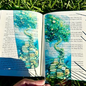 May include: A rectangular, transparent bookmark featuring a vibrant illustration of a large, leafy tree growing out of a stack of colorful books. The artwork displays shades of blue, green, and yellow, with intricate details of branches and foliage.