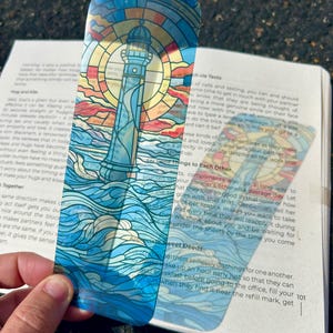 May include: A colorful bookmark featuring a lighthouse design. The lighthouse is depicted in a stained-glass style with blue, yellow, and red hues, set against a backdrop of ocean waves and a sunset sky. The bookmark is held over an open book.