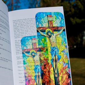 May include: A colorful bookmark featuring a stained-glass style depiction of Jesus on the cross. The artwork uses a vibrant palette of blues, yellows, and reds. The bookmark is held open in a book.