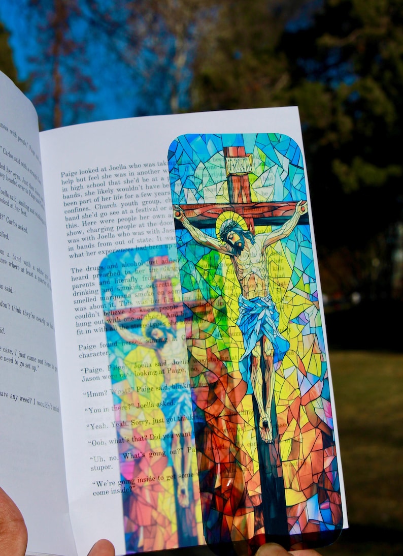 May include: A colorful bookmark featuring a stained-glass style depiction of Jesus on the cross. The artwork uses a vibrant palette of blues, yellows, and reds. The bookmark is held open in a book.