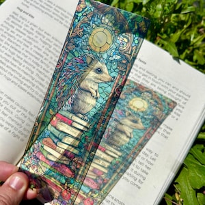 May include: A rectangular bookmark featuring a stained glass style illustration of a hedgehog perched atop a stack of colourful books. The design includes a circular sun motif at the top and intricate leaded glass patterns in shades of blue, green, and red.