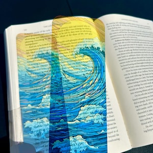May include: A clear, rectangular bookmark featuring a vibrant ocean wave design in shades of blue, white, and yellow. The bookmark is placed in an open book, with the wave illustration prominently displayed. A unique accessory for book lovers.