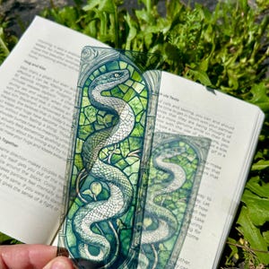 May include: A rectangular, semi-transparent bookmark featuring a detailed illustration of a coiled snake. The artwork mimics a stained-glass window design with shades of green, white, and black, framed by intricate, dark, swirling decorative borders.